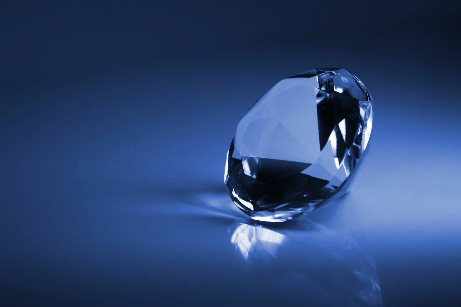 Ein geschliffener Diamant liegt auf einer blauen, reflektierenden Oberfläche und wirft einen Schatten. Die Facetten des Diamanten sind scharf und funkeln leicht im Licht.